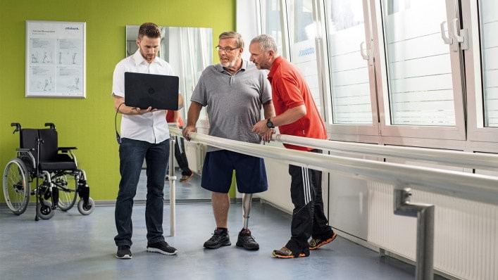 Patient mit Prothese steht mit Techniker und Therapeut in einem Anproberaum.